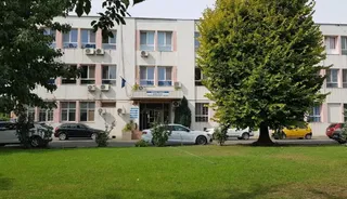 O clădire de trei etaje cu fațadă albă și roz este vizibilă în spatele unor copaci, cu mai multe mașini parcate în prim-plan.
