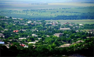 O vedere panoramică asupra unui sat cu case cu acoperișuri colorate înconjurate de vegetație abundentă și câmpuri deschise în fundal.