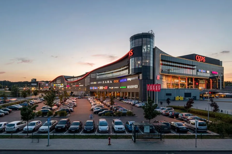 O clădire comercială modernă cu fațadă de sticlă și panouri luminoase este situată în spatele unui parcări plin de mașini colorate sub un cer la apus.