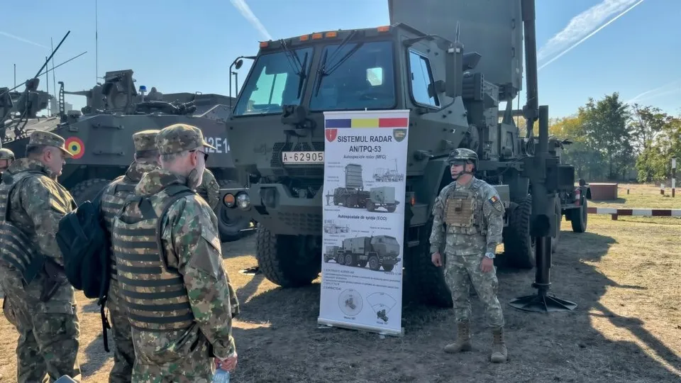 Soldați în uniforme de camuflaj stau lângă un vehicul militar mare și un panou informativ vertical pe un teren cu iarbă.