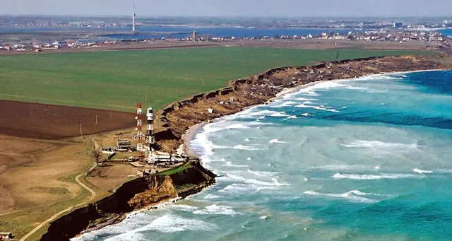 O vedere aeriană a unei coaste cu o faleză stâncoasă, un câmp verde întins și apele mării cu valuri albe spumante.