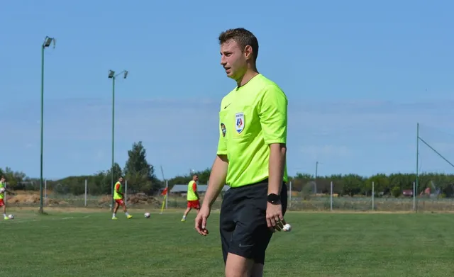 Un bărbat într-un tricou galben și pantaloni scurți negri stă pe un teren de fotbal verde sub un cer senin.