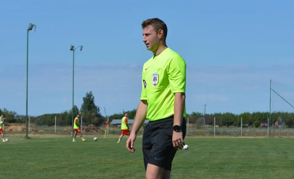Un bărbat într-un tricou galben și pantaloni scurți negri stă pe un teren de fotbal verde sub un cer senin.