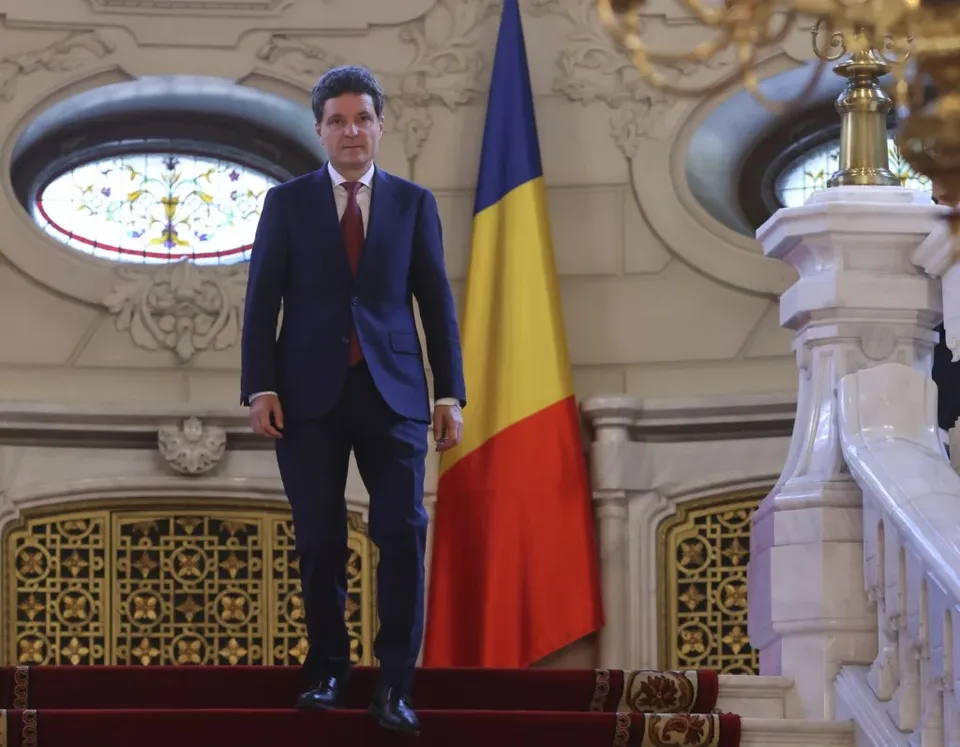 Un bărbat într-un costum albastru și cravată roșie coboară treptele unei scări interioare lângă un steag tricolor.