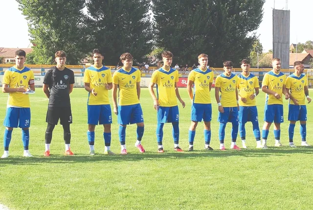 O echipă de fotbaliști stă în rând pe un teren de gazon verde, purtând echipamente sportive galbene și albastre sau negre.