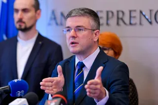 Un bărbat cu ochelari și costum în bleu arată cu mâinile deschise în timp ce vorbește în fața unor microfoane.