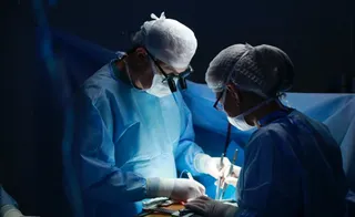 Doi medici în halate și căciuli sterile de operație execută o procedură chirurgicală sub lumină intensă într-un cadru întunecat.
