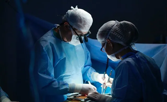 Doi medici în halate și căciuli sterile de operație execută o procedură chirurgicală sub lumină intensă într-un cadru întunecat.