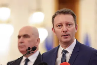 Un bărbat cu cămașă albă, cravată cu puncte și sacou închis la culoare vorbește în fața unor microfoane, având un alt bărbat în fundal.