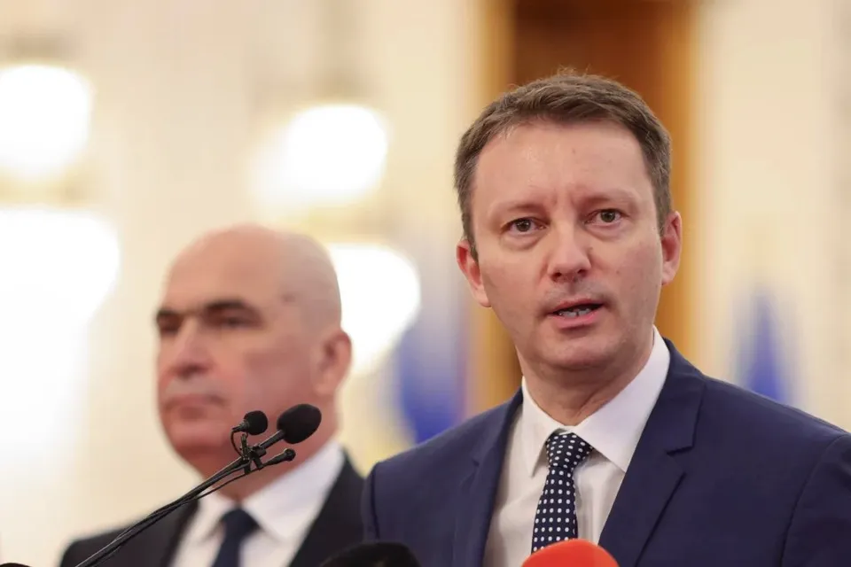 Un bărbat cu cămașă albă, cravată cu puncte și sacou închis la culoare vorbește în fața unor microfoane, având un alt bărbat în fundal.