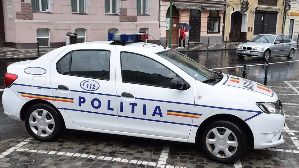 O mașină de poliție albă cu inscripții albastre și galbene este parcată pe un drum umed lângă clădiri urbane.