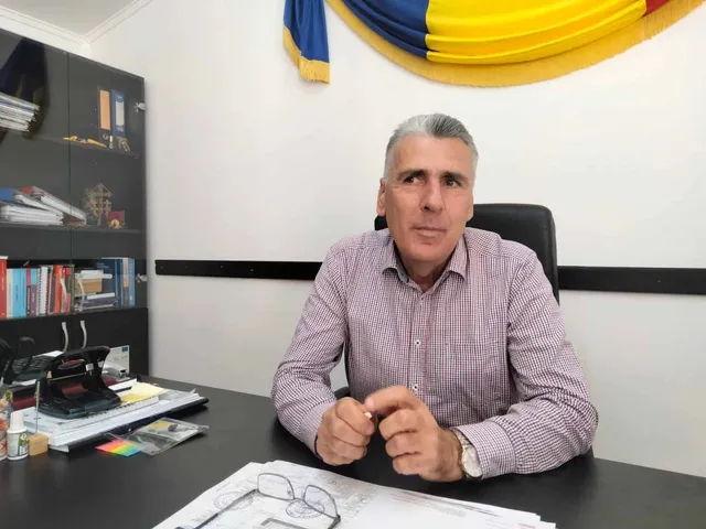 Un bărbat cu părul cărunt și cămașă roz stă la un birou lângă un raft cu cărți și un drapeau.