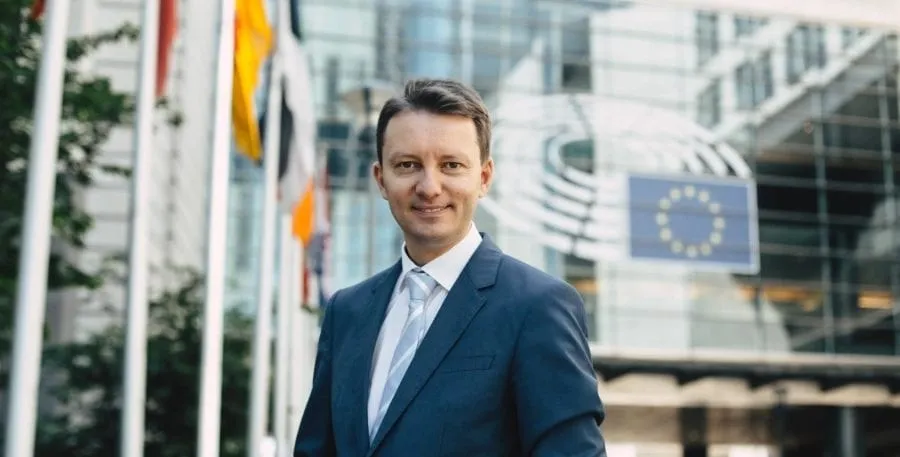 Un bărbat cu părul scurt, purtând un costum albastru și o cămașă albă cu cravată, stă în fața unei clădiri cu un logo și steaguri.