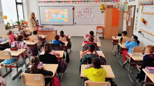 O femeie stă în fața unui ecran de proiecție într-o sală de clasă în timp ce copiii stau la bănci întoși spre ea.