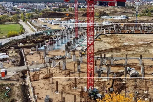 O vedere de sus prezintă un șantier cu piloni de beton, excavări, utilaje de construcții și o macararie roșie înaltă.