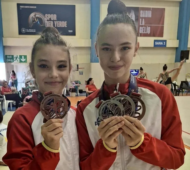 Două tinere purtând costume roșii și albe țin în mâini medalii mari cu fundal roșu și negru într-o sală de sport.