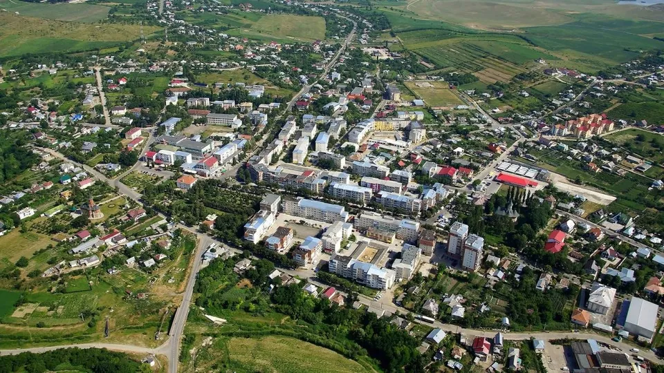 O vedere aeriană prezintă o zonă urbană cu clădiri diverse, drumuri și terenuri verzi înconjurate de câmpuri agricole.