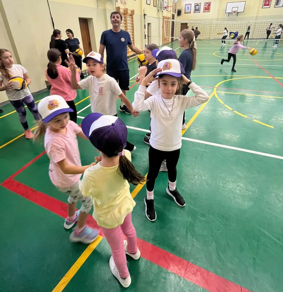 Copii cu veselii, capace de baseball și echipamente sportive, stând pe un parchet de sală de gimnastică cu linii colorate.