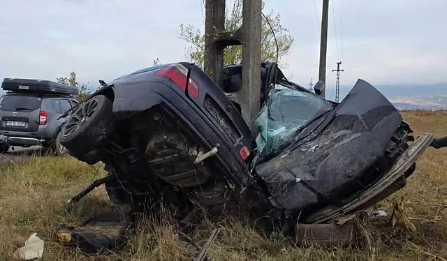 Un autoturism negru este răsturnat și lovit de un stâlp de beton într-un câmp cu iarbă uscată sub un cer înnorat.