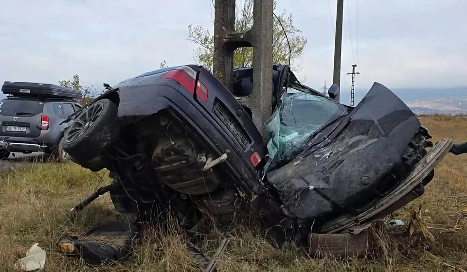 Un autoturism negru este răsturnat și lovit de un stâlp de beton într-un câmp cu iarbă uscată sub un cer înnorat.