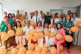 Un grup de persoane în haine albe și colorate stau grupate în fața unui perete alb, ținând numeroase baloane portocalii.