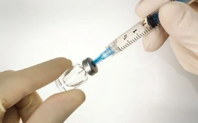 Mâini încăpățânate cu mănuși albe țin o seringă cu lichid albastru și un flacon de sticlă transparent cu dop metalic.
