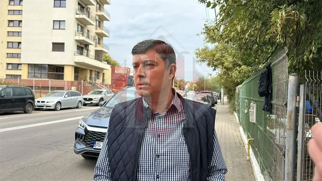 Un bărbat cu părul scurt și cămașă în croit sub un vest negru stă pe un trotuar lângă un gard și mașini parcate.