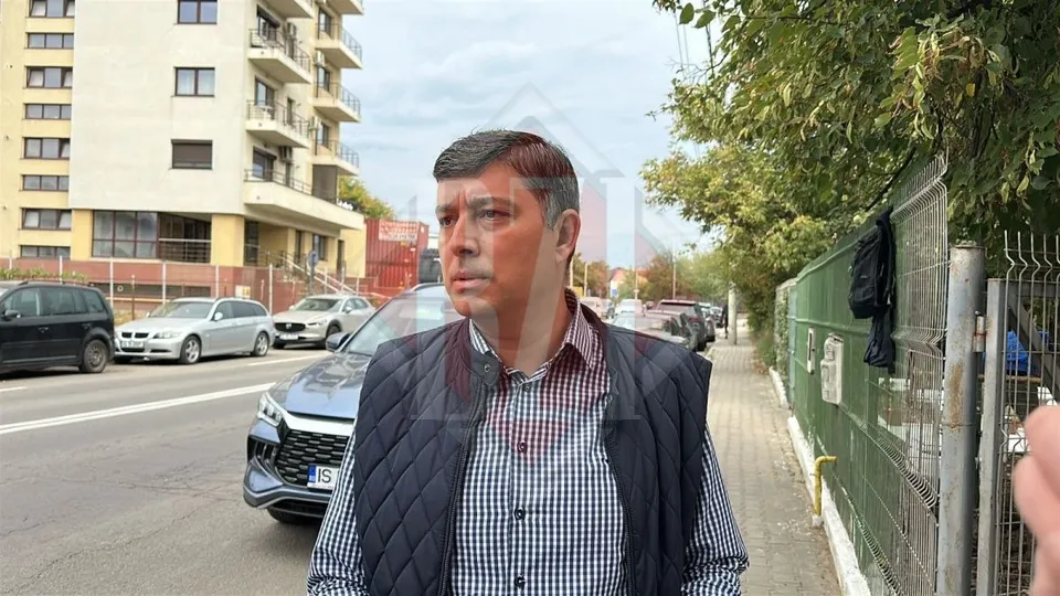 Un bărbat cu părul scurt și cămașă în croit sub un vest negru stă pe un trotuar lângă un gard și mașini parcate.