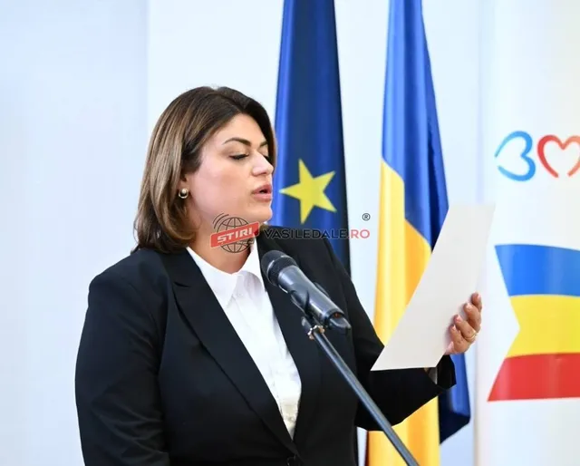 O femeie cu părul castaniu, purtând un sacou negru și o cămașă albă, ține o foaie de hârtie lângă un microfon.