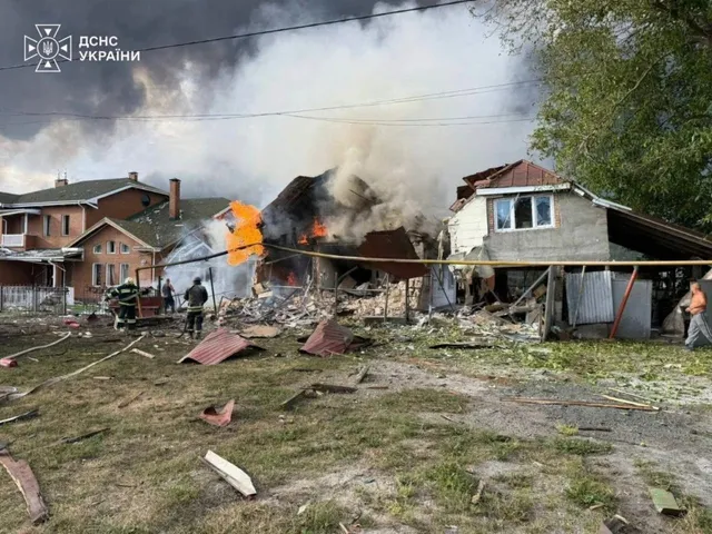 Focuri de flacără și fum dens ies dintr-o clădire distrusă, în timp ce personal în uniforme de protecție se află în fața structurii.