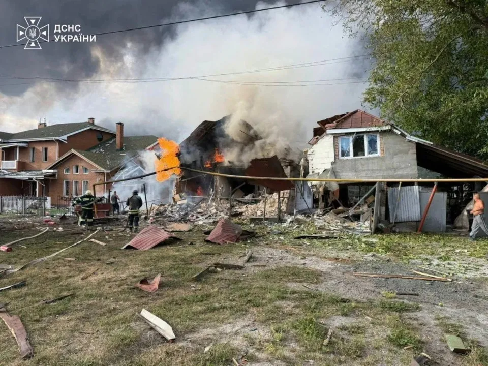 Focuri de flacără și fum dens ies dintr-o clădire distrusă, în timp ce personal în uniforme de protecție se află în fața structurii.