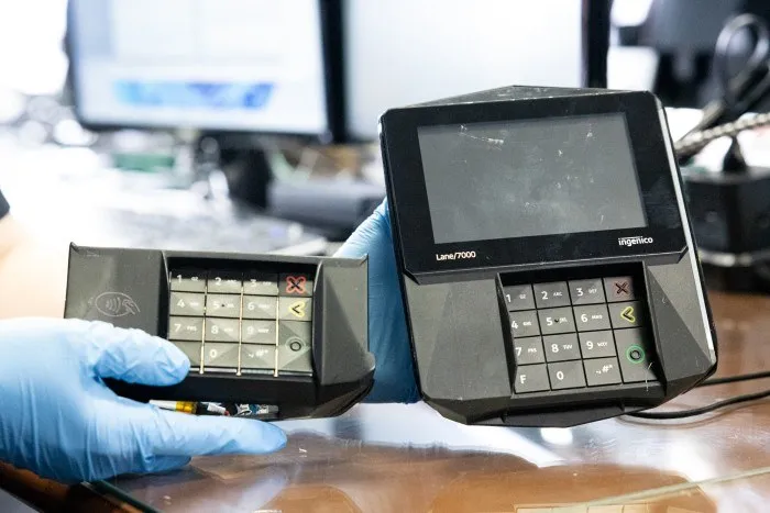 O persoană cu mănuși albastre de protecție ține un dispozitiv electronic de plată negru lângă un alt terminal de carduri similar pe o masă.