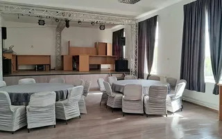 O sală cu mese rotunde acoperite cu fețe de masă gri și scaune cu huse albe, situată în fața unei scene cu structură metalică.