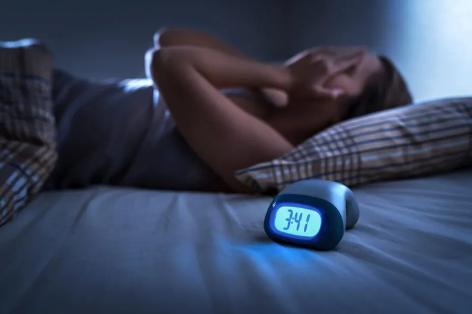 O persoană stă întinsă pe o pat cu mâinile la față, lângă un ceas digital care afișează cifra 3:41.