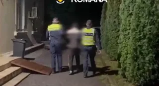 Trei persoane, dintre care două poartă veste galbene, stau pe un drum lângă un gard de vegetație densă pe timp de noapte.