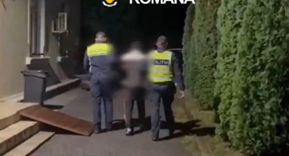 Trei persoane, dintre care două poartă veste galbene, stau pe un drum lângă un gard de vegetație densă pe timp de noapte.