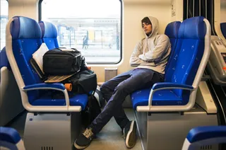 Un bărbat cu hanorac gri stă așezat cu brațele încrucișate pe un scaun albastru dintr-un tren, lângă o fereastră mare.
