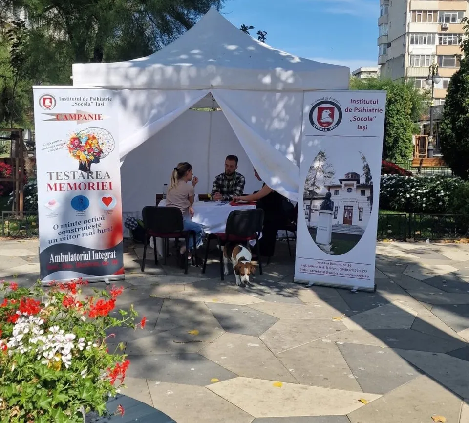 O femeie și un bărbat stau la o masă sub un cort alb, lângă un panou informativ cu text și imagini.