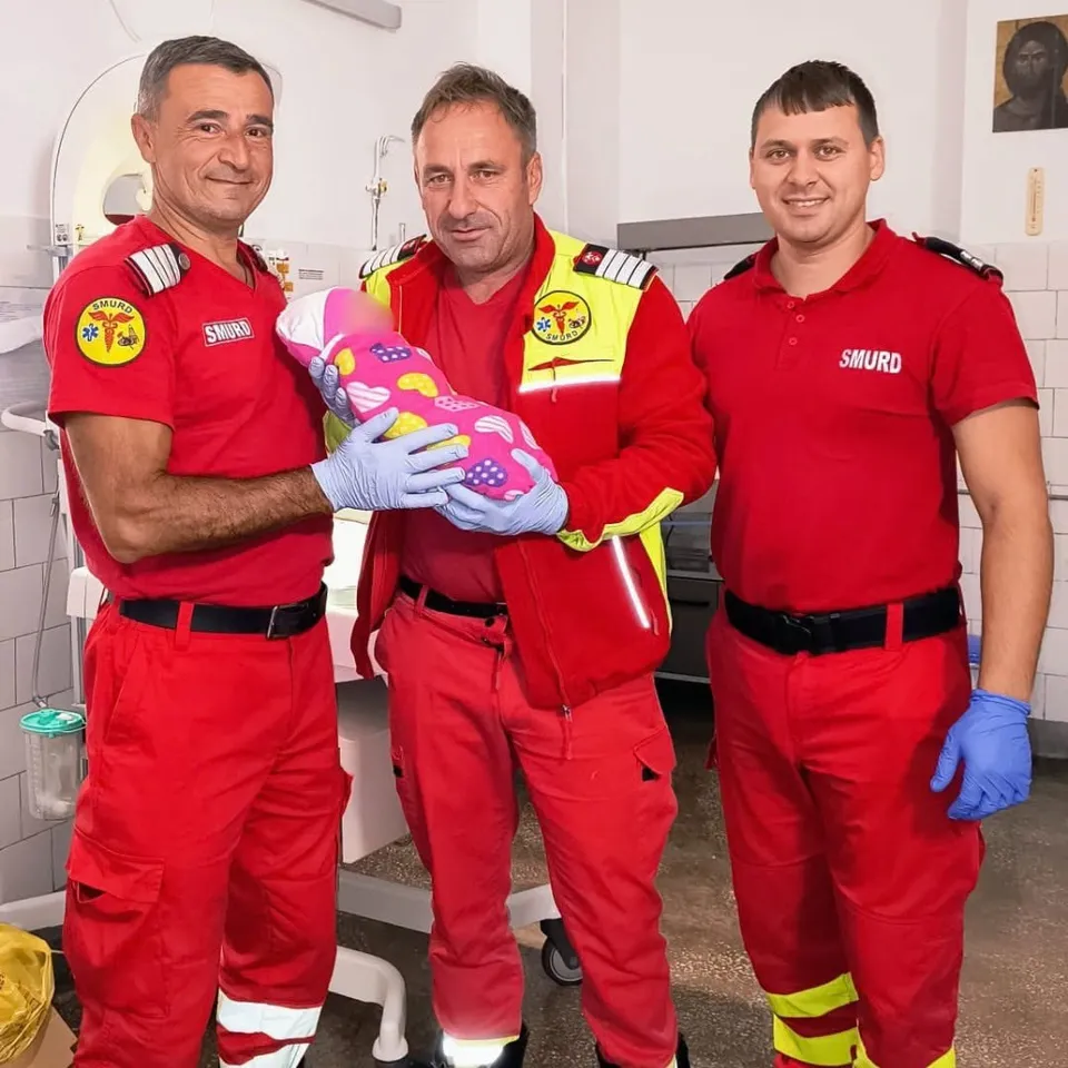 Trei bărbați în uniforme roșii de salvamontaj țin împreună un nou-născut învelit într-o pătură roz cu modele colorate într-un mediu medical.