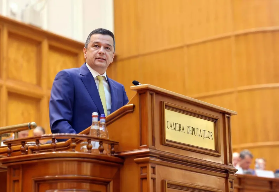 Un bărbat într-un costum albastru și cravată galbenă stă în spatele unui podium de lemn pe care scrie Camera Deputaților.