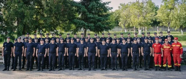 Un grup de oameni în uniforme de serviciu, unele albastre și altele roșii, stau aliniați în rânduri pe un teren deschis lângă copaci.