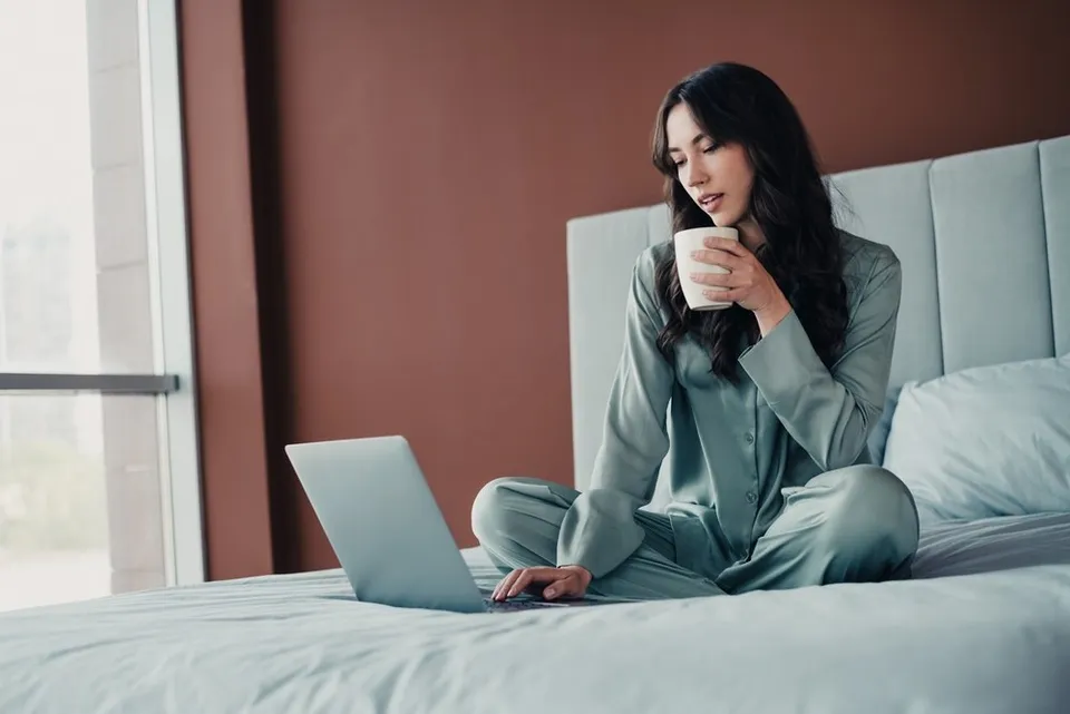O femeie cu părul lung stă încrucișată pe un pat, purtând o pijama de satin și ținând o cană în timp ce privește un laptop deschis.