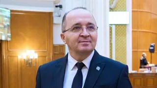 Un bărbat cu ochelari și cravată neagră poartă un sacou închis la culoare în fața unui fundal cu uși de lemn și panouri de perete.