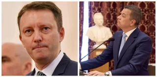 Un bărbat cu părul scurt și costum încadrează planul din față, în timp ce un alt bărbat în costum stă în fața unui microfon.
