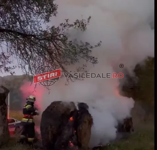 Un pompier cu echipament de protecție și cască se află lângă un balot de fân care arde și scoate fum dens.