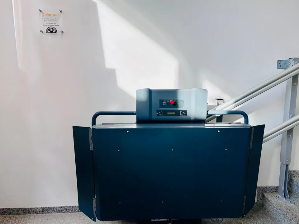 O platformă hidraulică de culoare închisă este instalată lângă o rampă cu balustradă metalică într-un spațiu cu pereți albi.