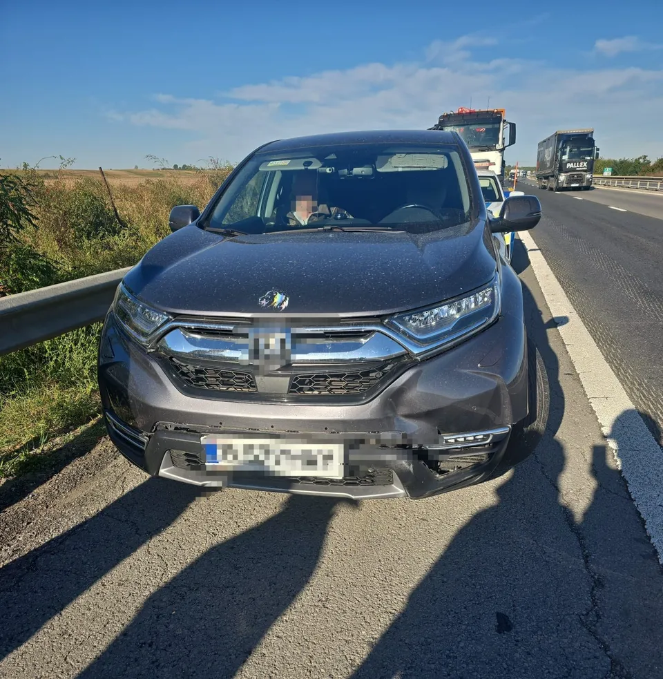 Un autoturism gri cu partea frontală avariată este oprit pe marginea unei autostrăzi lângă un parapet metalic sub un cer senin.
