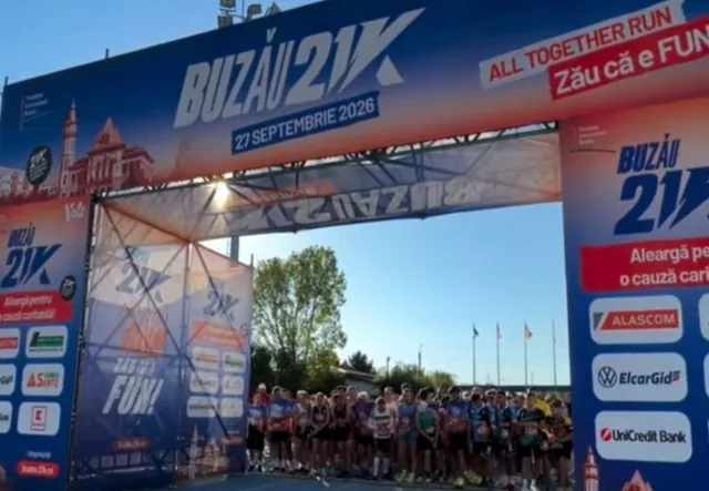 Un grup de sportivi stă în rând în spatele unei porți de start pentru o cursă de alergare sub un cer senin.