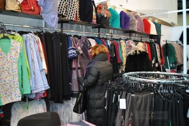 O persoană cu părul roșcat, purtând o haină neagră lungă, privește spre haine colorate suspendate pe umerașe într-un magazin sau târg.