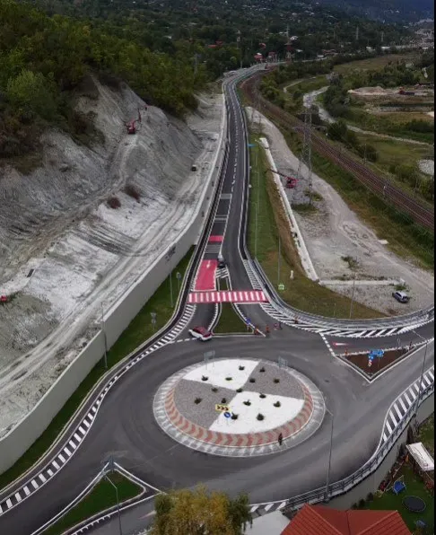 O vedere aeriană prezintă o intersecție de tip rondel cu asfalt nou, marcaje rutiere albe și linii de siguranță, situată lângă un versant de deal.